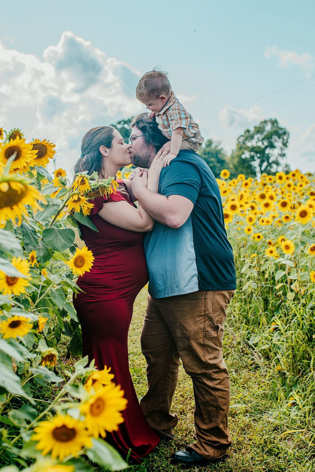 Couple kissing while holding a child in a sunflower field under a bright sky