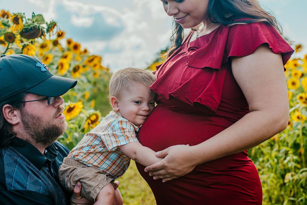 Pregnant woman in red dress holding toddler beside bearded man in cap in a sunflower field