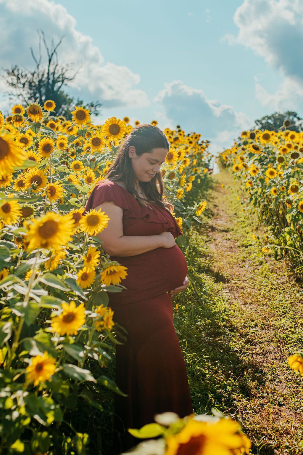 Pregnant woman in a burgundy dress standing in a sunflower field under a bright blue sky