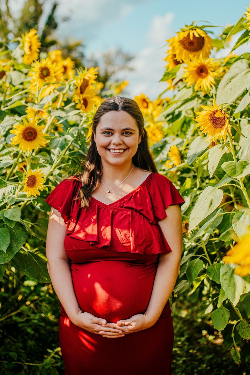 Woman in a red dress smiling in a sunflower field, hands clasped.