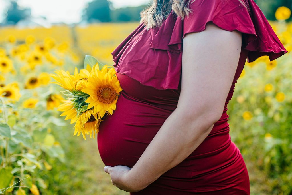 Pregnant person in a red dress standing in a sunflower field, holding sunflowers