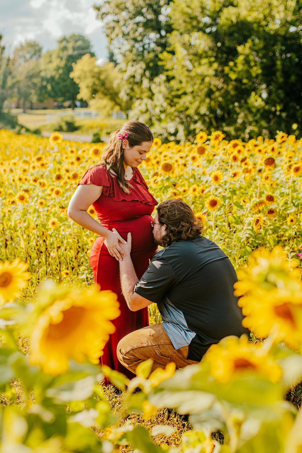 Person kneeling in a sunflower field adjusting a pregnant woman’s red dress outdoors