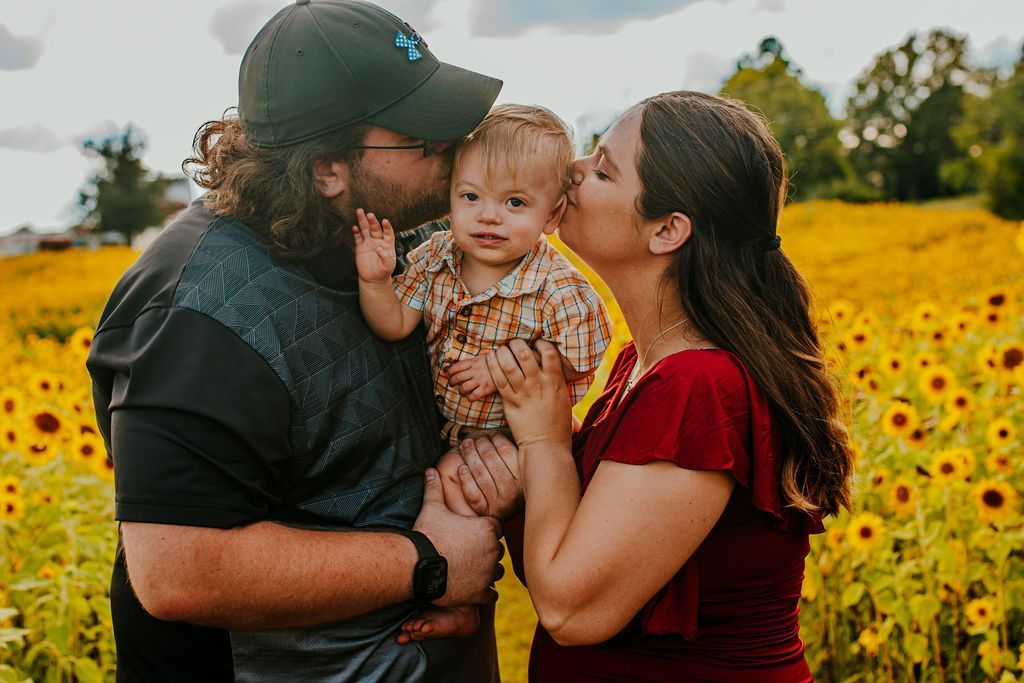Family of three embracing in a sunflower field, with parents kissing a toddler between them.