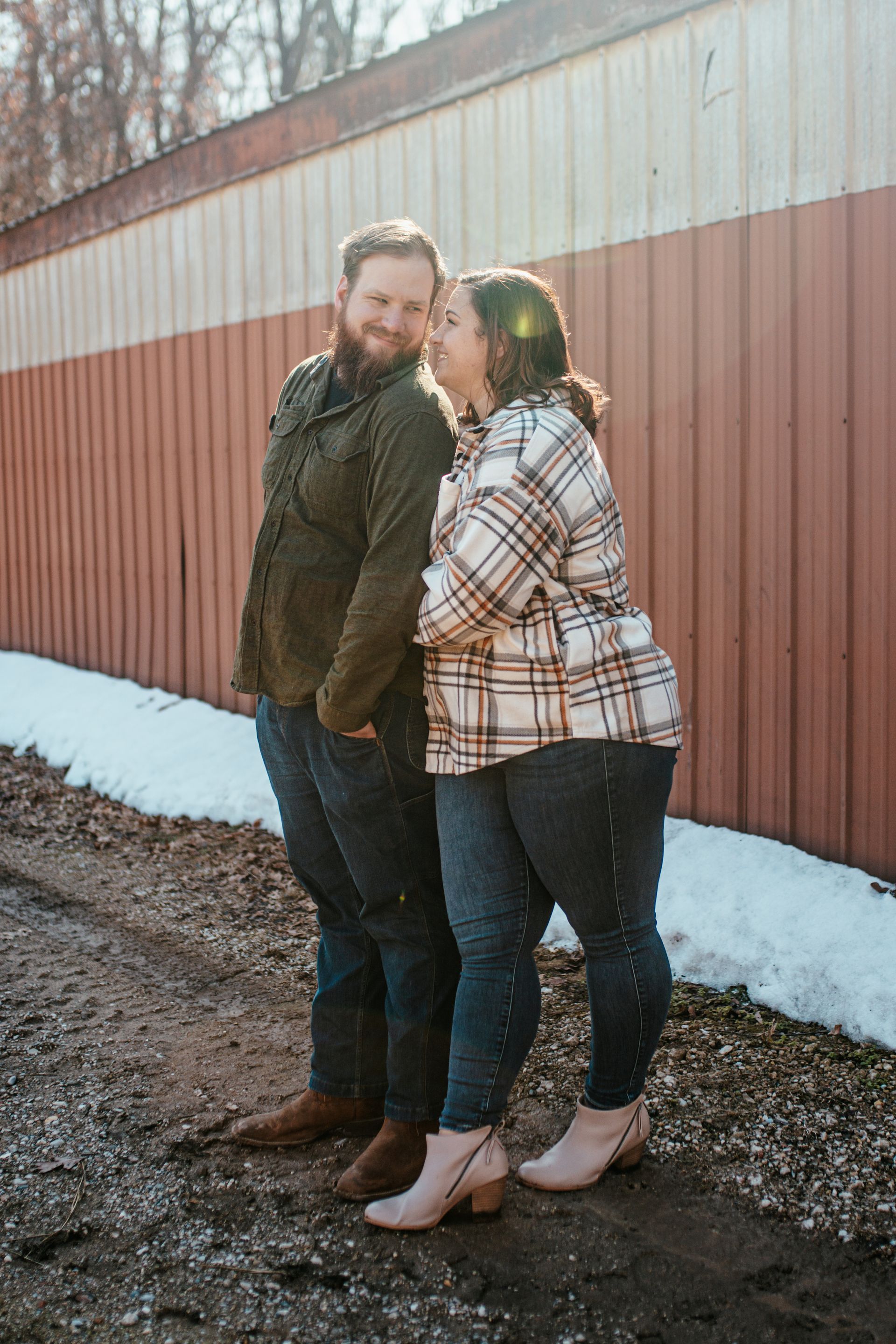 Two people standing together outdoors by a red barn wall, smiling in plaid jackets.