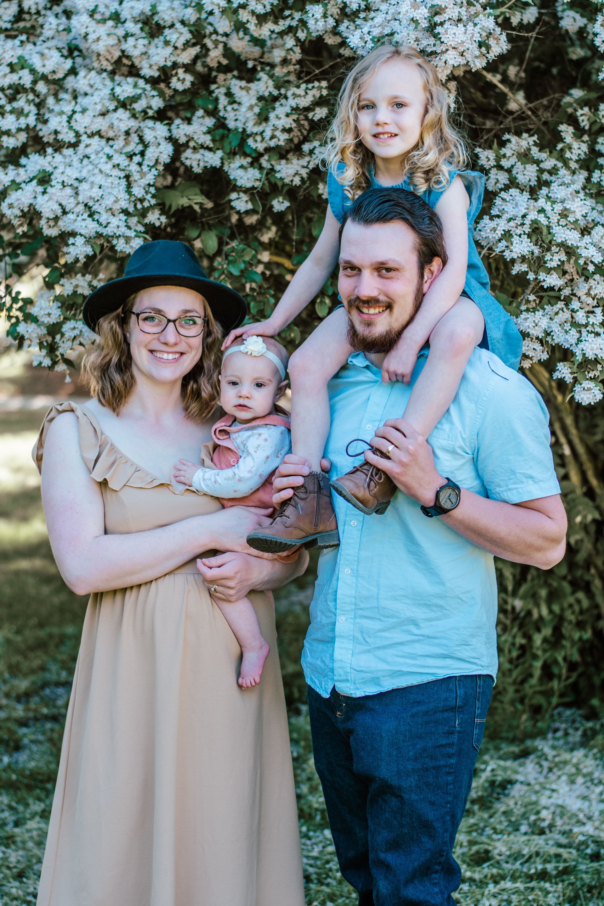 Family of four posing outdoors in front of white flowering bushes, with one child on each parent and smiling.