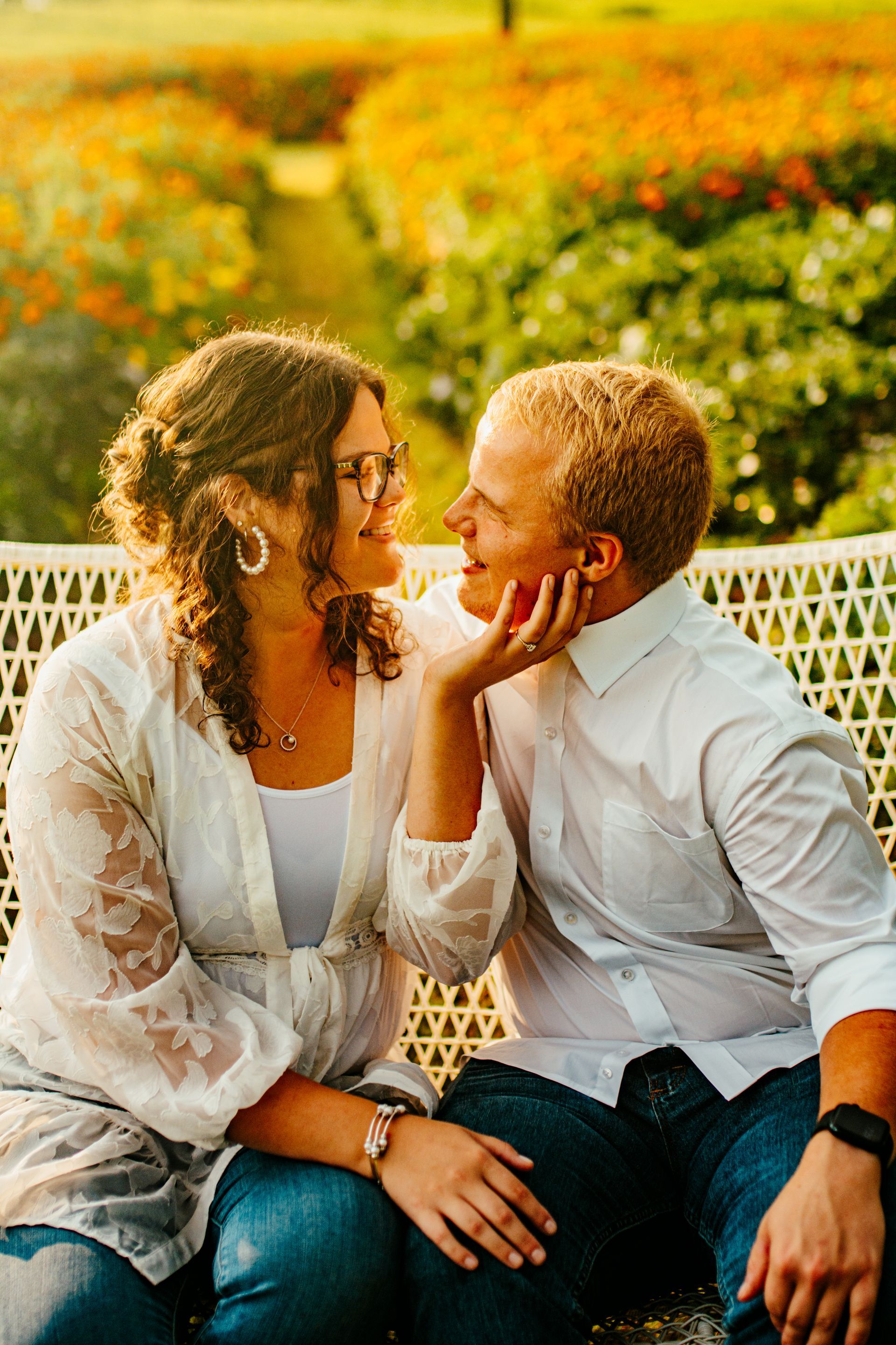 Two people sitting on a bench in a sunlit flower garden, smiling and touching faces.
