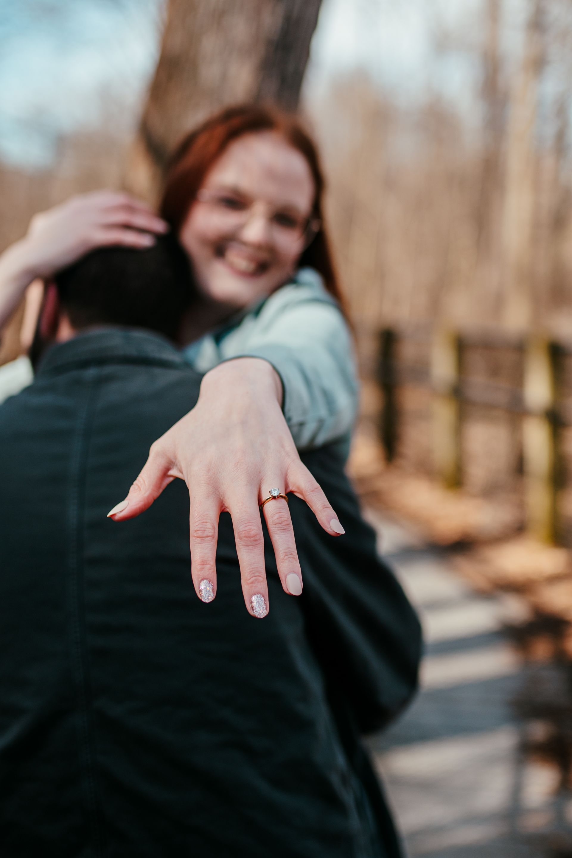 Woman with engagement ring reaching toward camera as partner hugs her on a wooded path