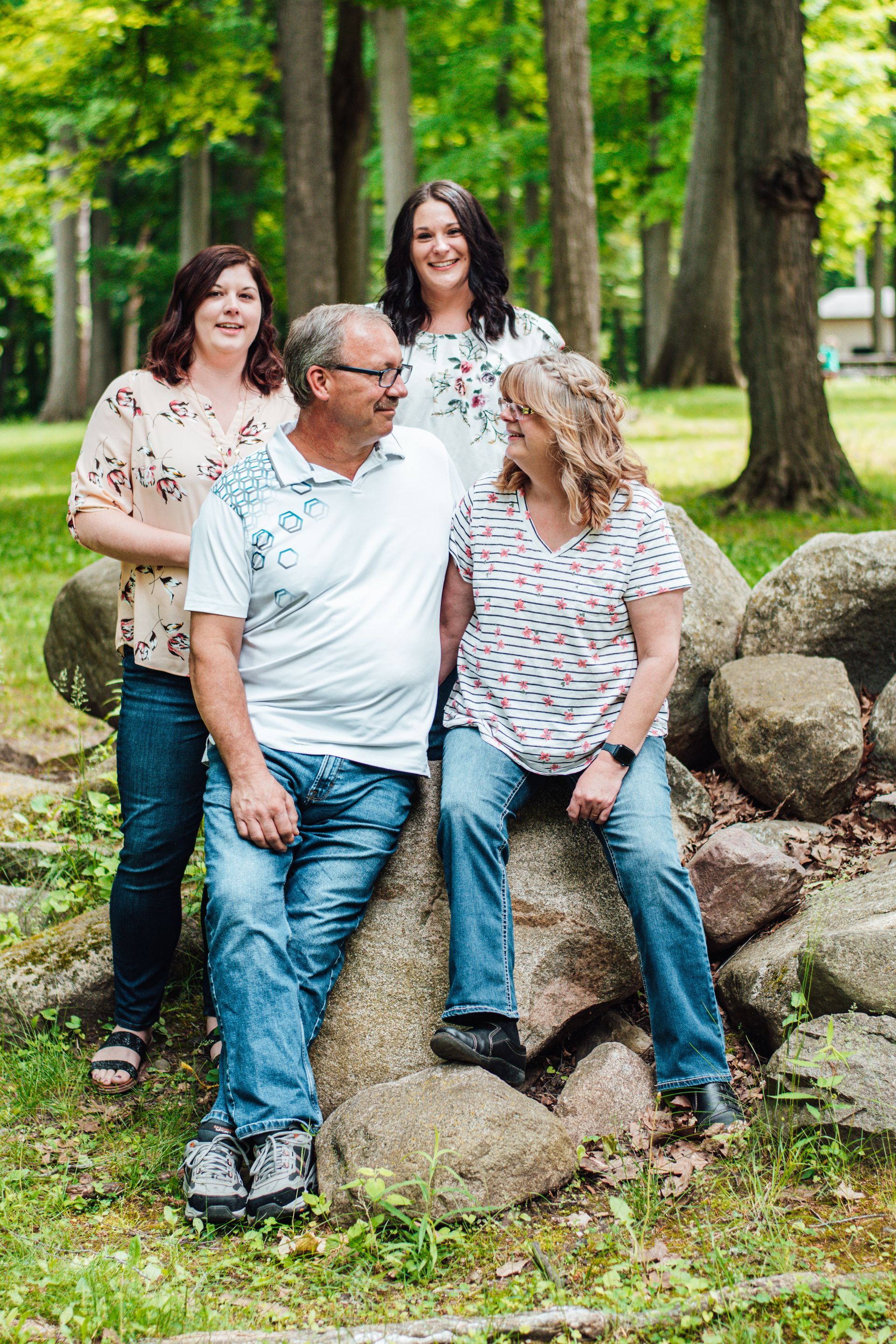 Four people pose outdoors among rocks and trees, with one seated on a stone and three standing behind.