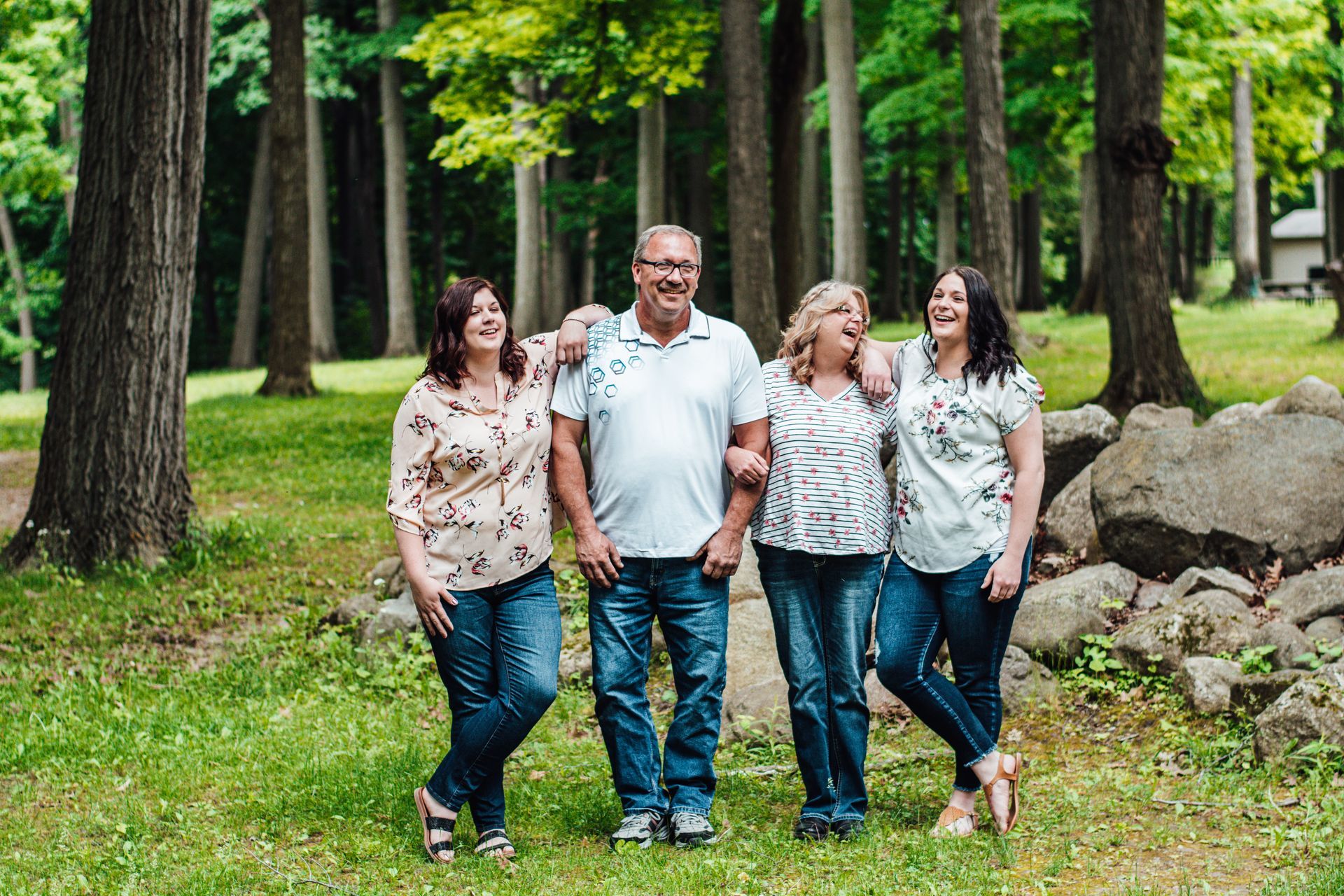 Four people walk together through a green park with trees and rocks, smiling at the camera.