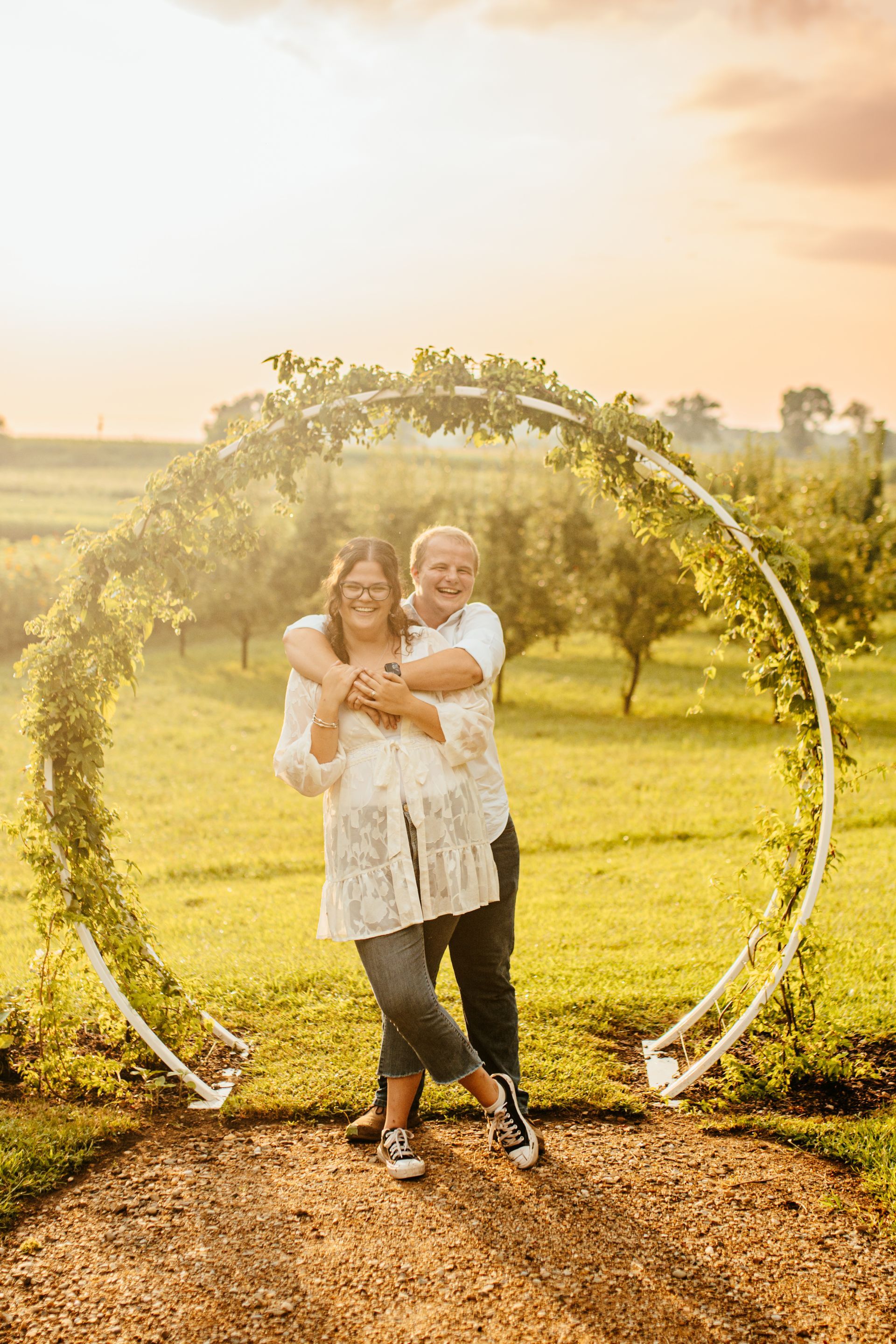Two people smiling in a floral arch in a sunlit field at sunset