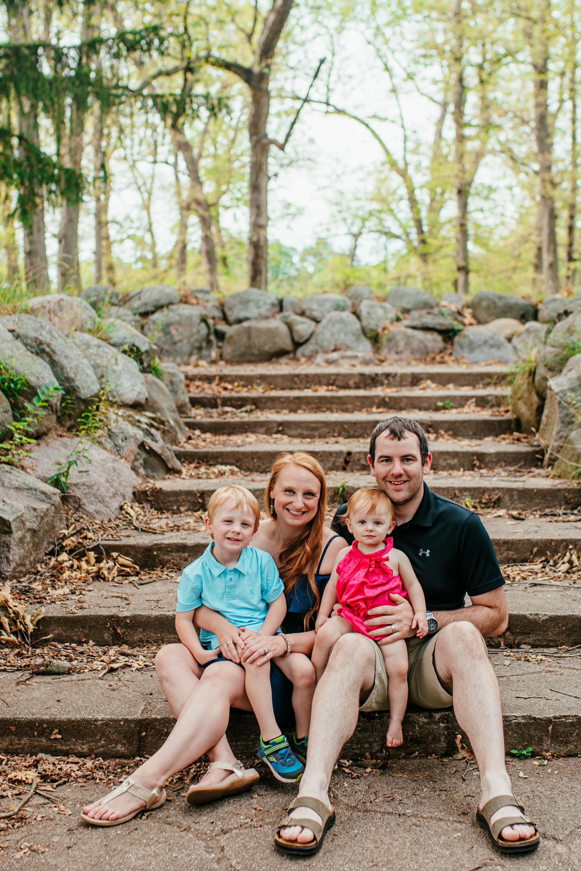 Family of four sitting on stone steps in a wooded park, smiling at the camera.