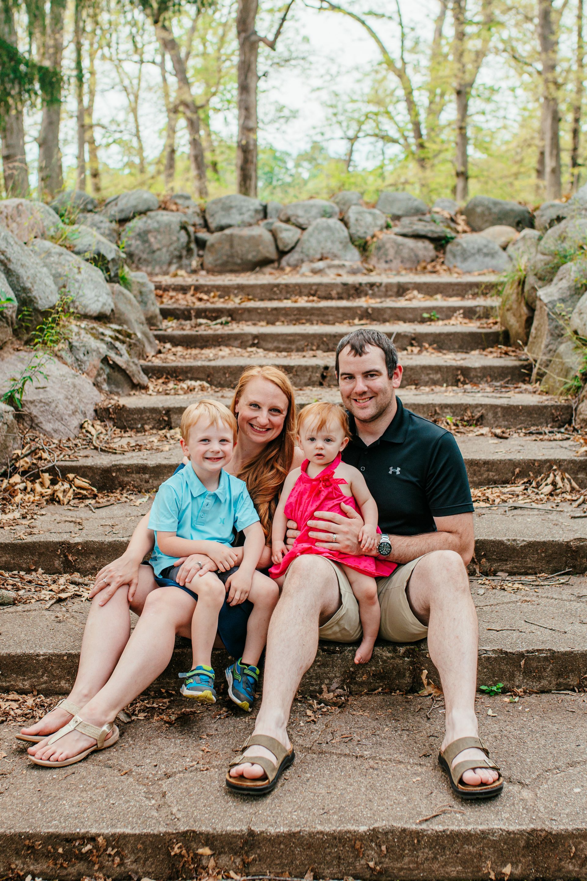 Family sitting on outdoor stone steps in a wooded park, smiling at the camera.