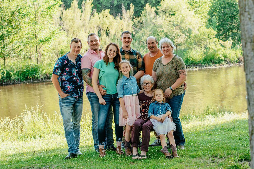 Group of seven people posing outdoors by a pond on a sunny day, with trees and grass in the background