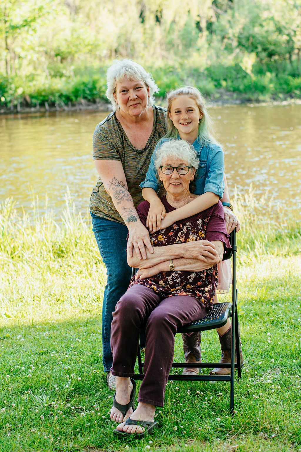 Three women posing outdoors by a pond, smiling and embracing in the grass