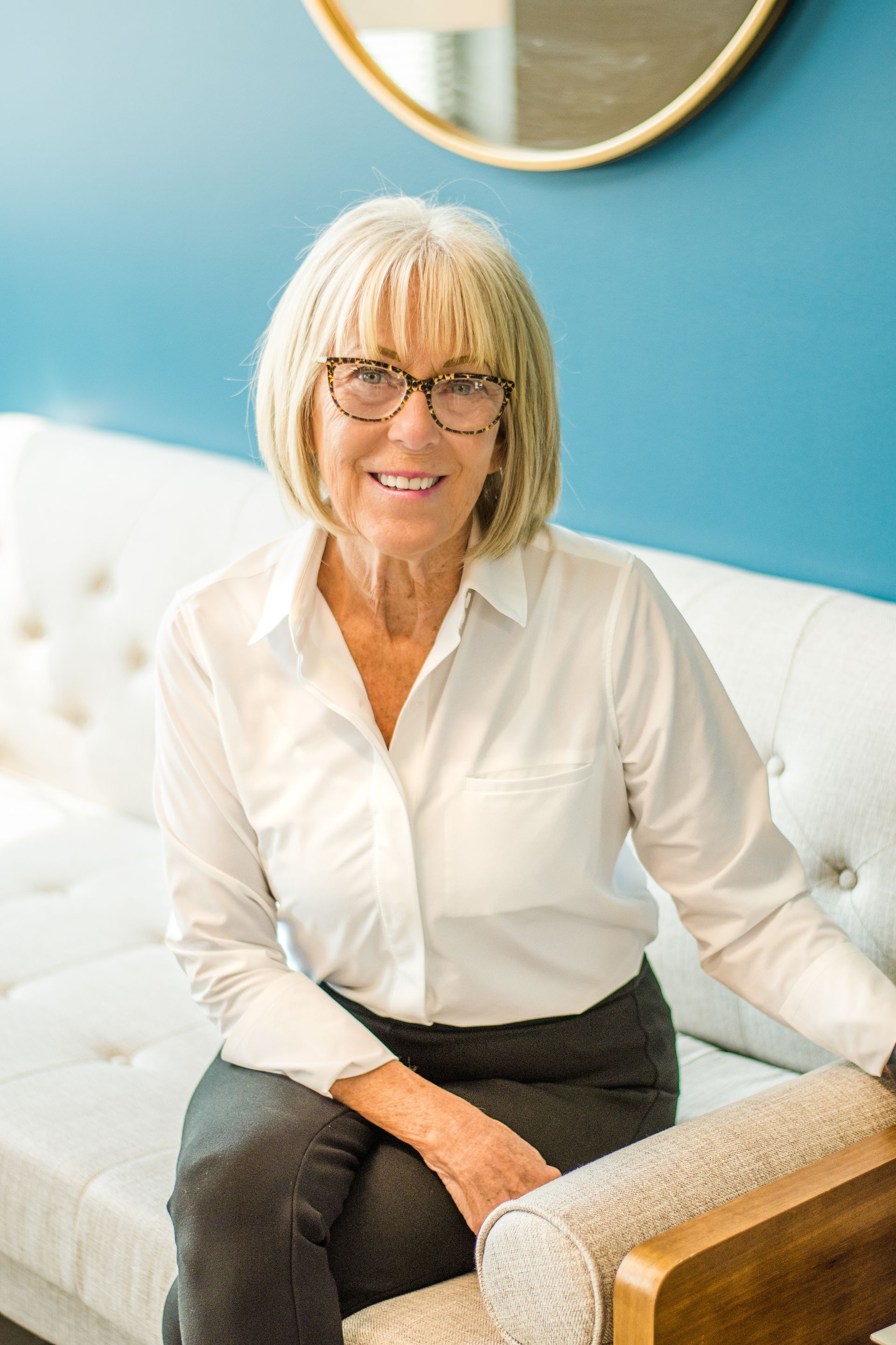Smiling woman seated on a white tufted sofa beside a turquoise wall and round mirror.