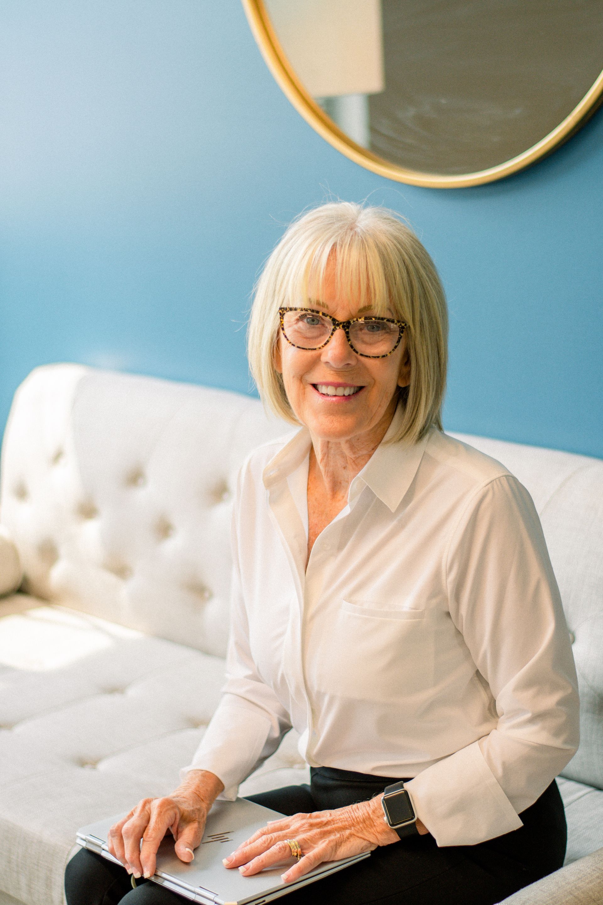 Woman sitting on a white tufted sofa beside a blue wall and round mirror, smiling at the camera.