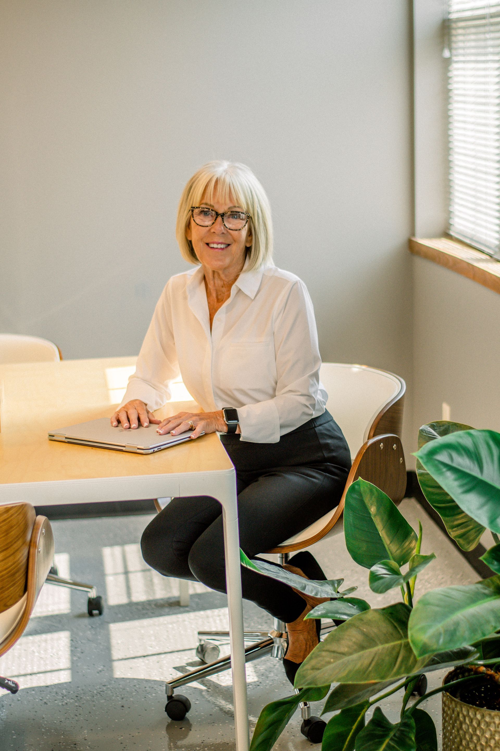 Smiling woman seated at a table in a bright office with plants and a window blind