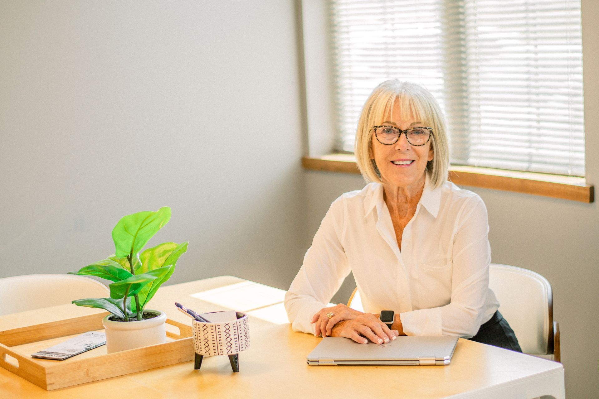 Smiling woman at a desk in a bright office, with a potted plant and window blinds behind her