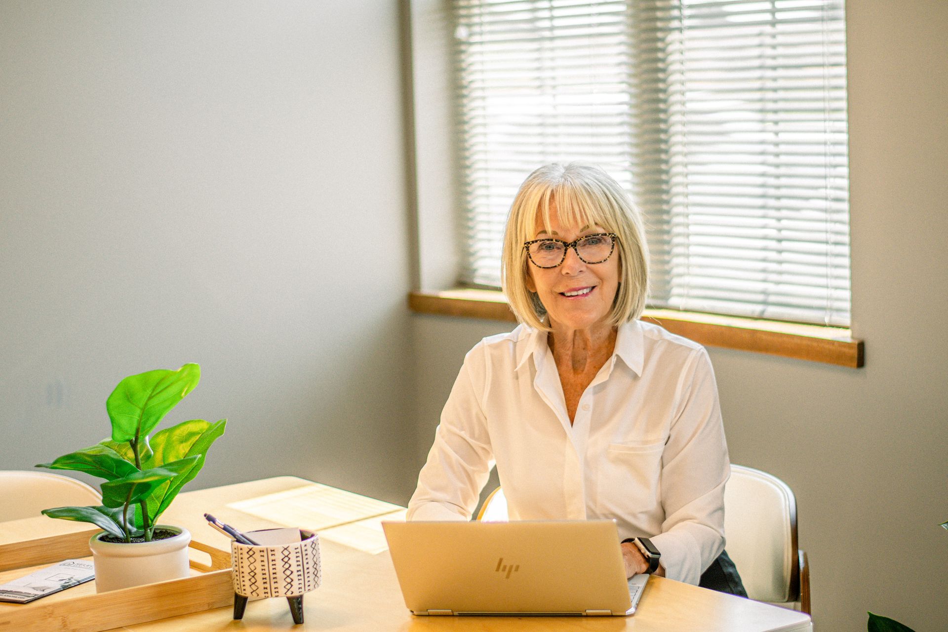Woman seated at a desk with a laptop in a bright office, smiling near a window and potted plant