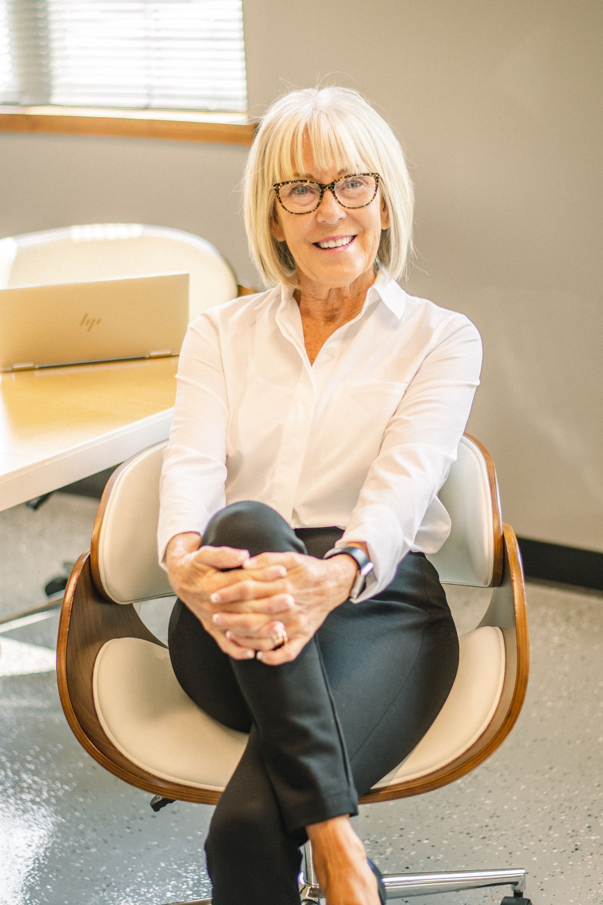 Smiling woman in white blouse seated in an office chair beside a desk, hands clasped