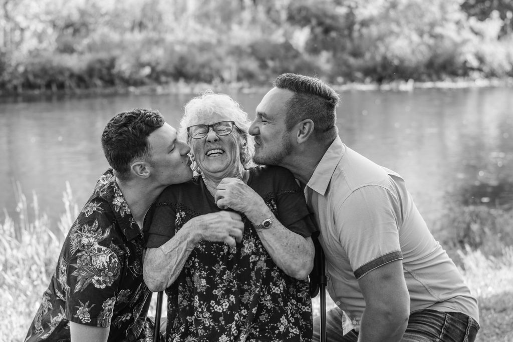 Two men kissing an elderly woman on both cheeks outdoors by a lake