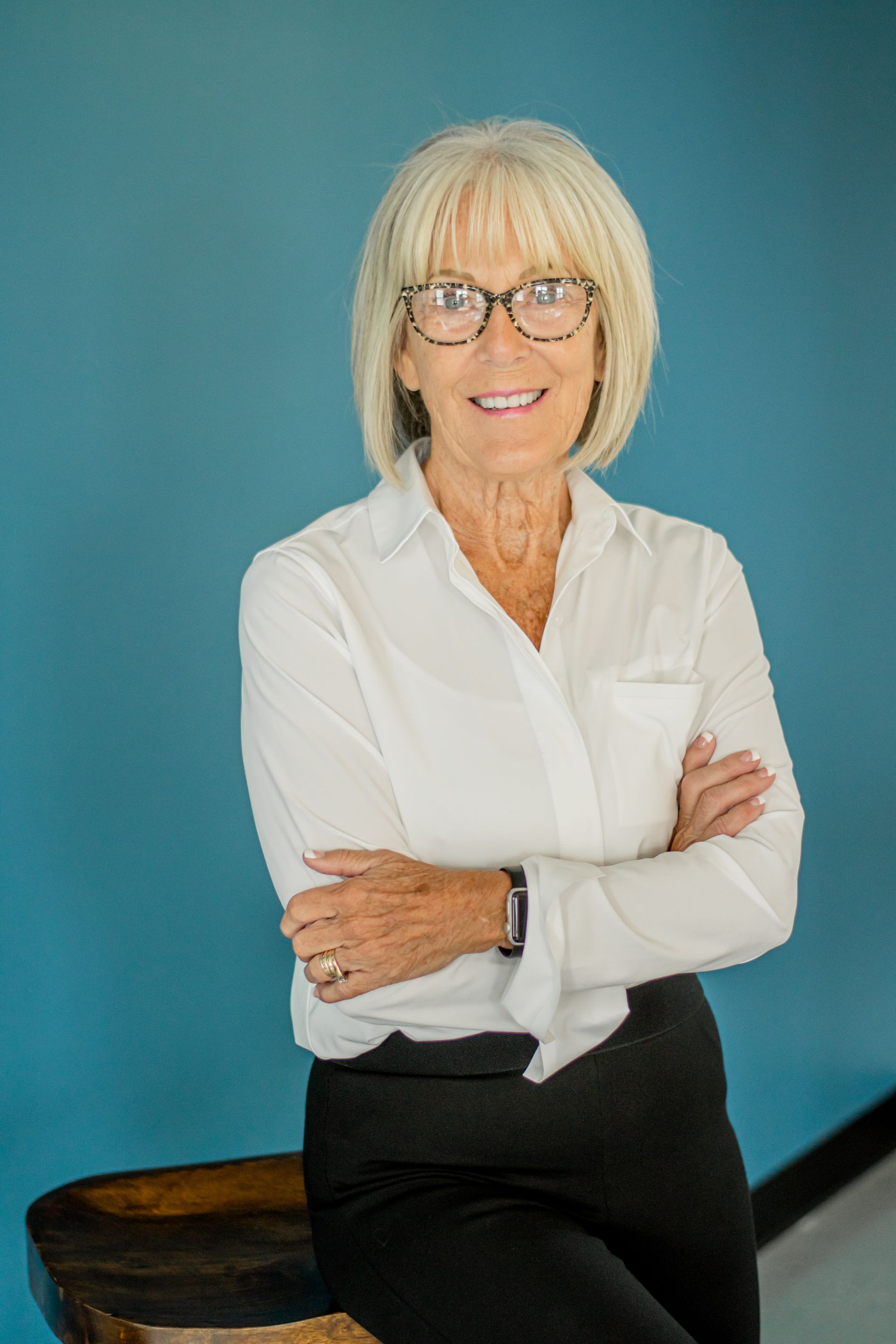 Portrait of a smiling woman with glasses, white blouse, and black pants, standing against a teal background.