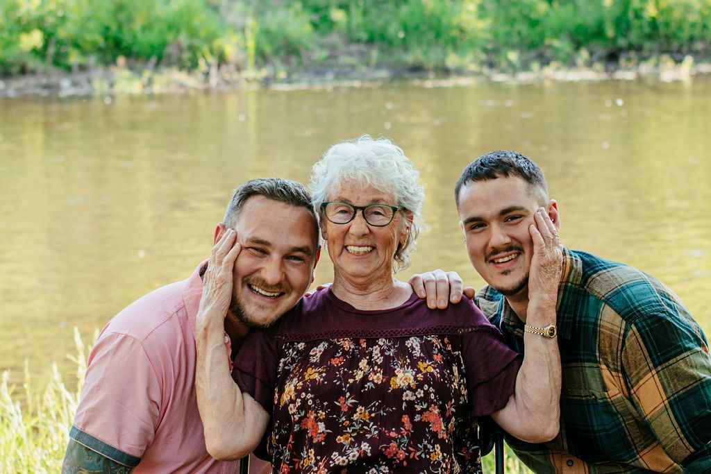 Three smiling people outdoors by a lake, leaning close together with arms around each other.