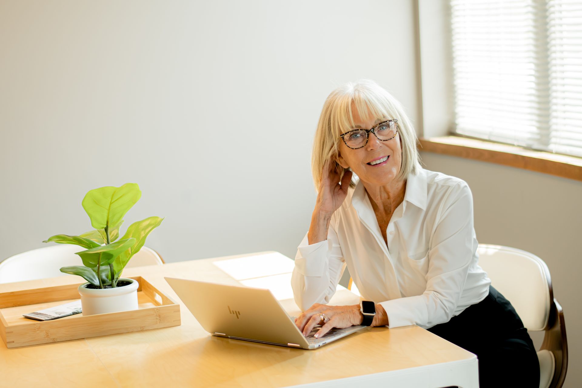 Woman working on a laptop at a sunlit table with a potted plant nearby