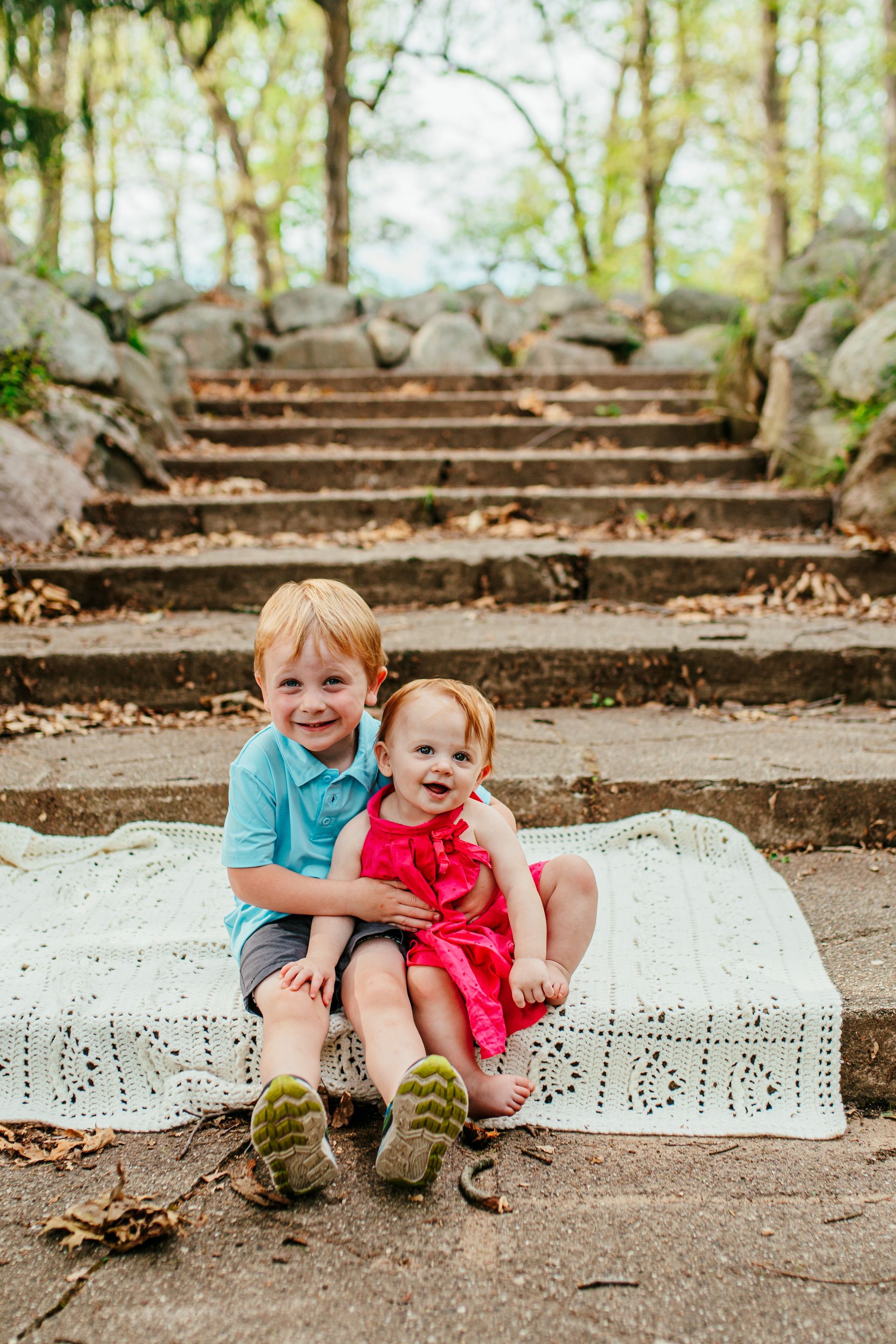 Two young children sitting on a white blanket on outdoor stone steps, smiling in a wooded area.
