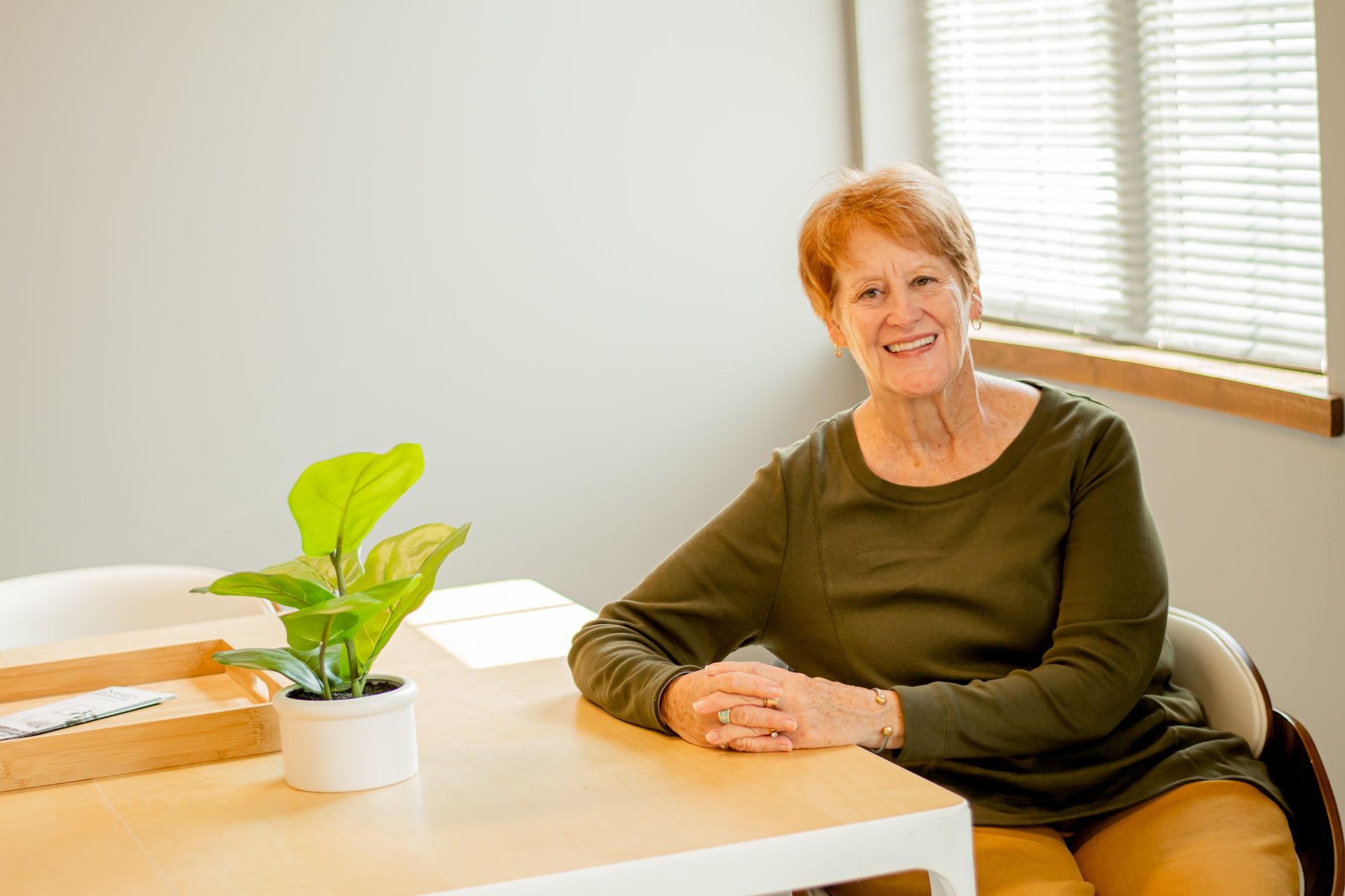 Woman seated at a table in a bright room, smiling beside a potted plant and window blinds