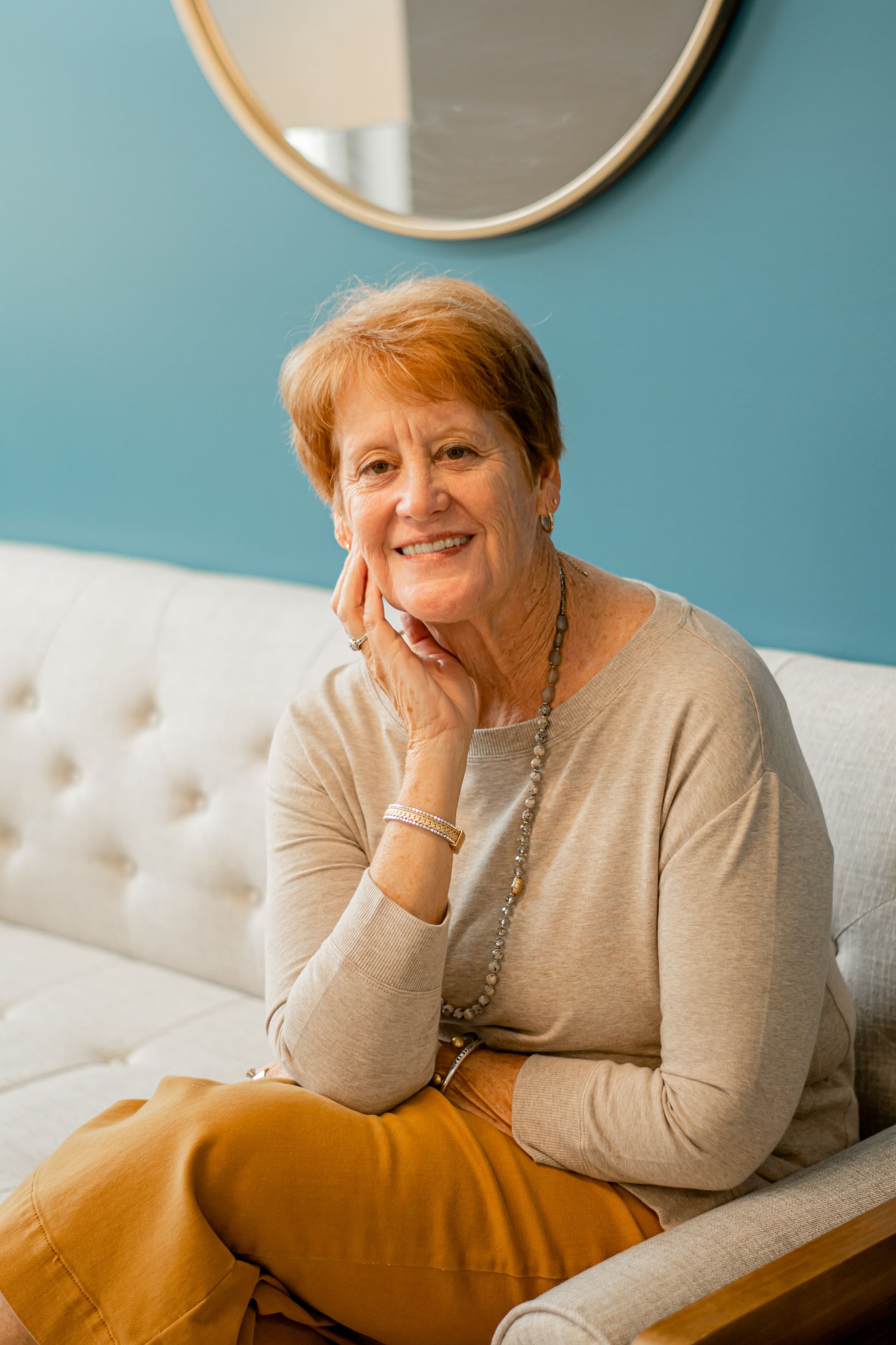 Smiling woman seated on a white couch against a teal wall, hand resting on her chin.