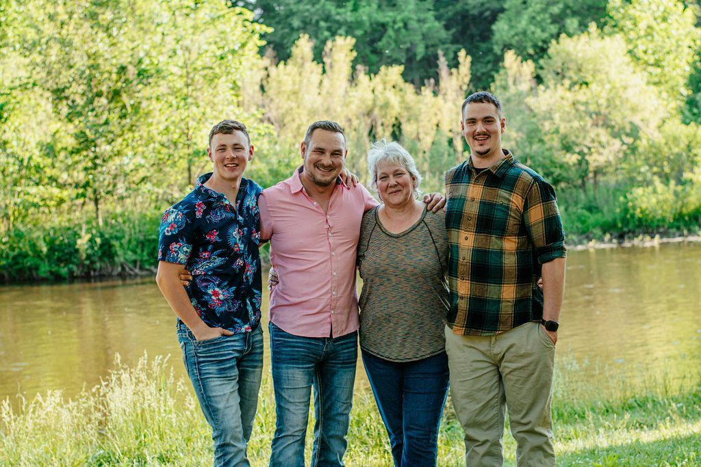 Four people standing together outdoors in front of a river and green trees, smiling at the camera.