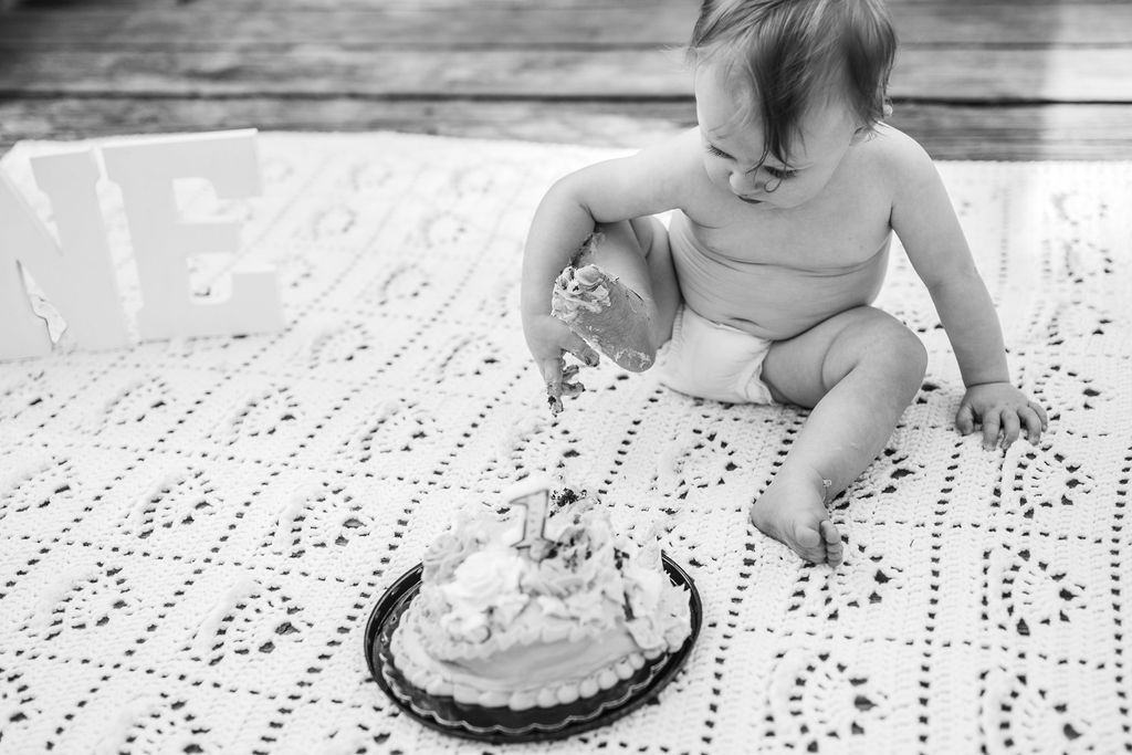 Toddler sitting on a blanket, reaching toward a cake on a plate, with a large “ONE” decoration nearby.