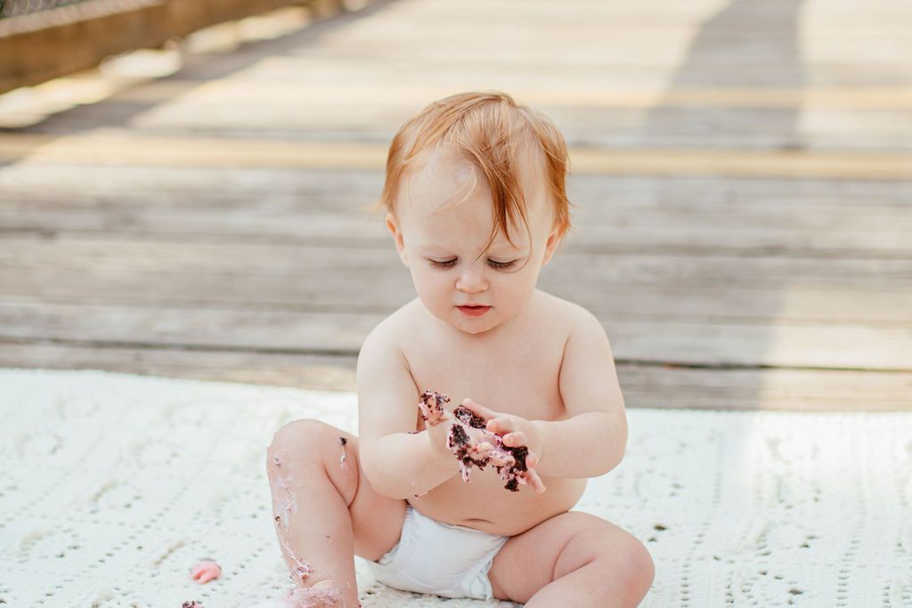 Toddler sitting on a light rug, looking down at their hands in a bright room
