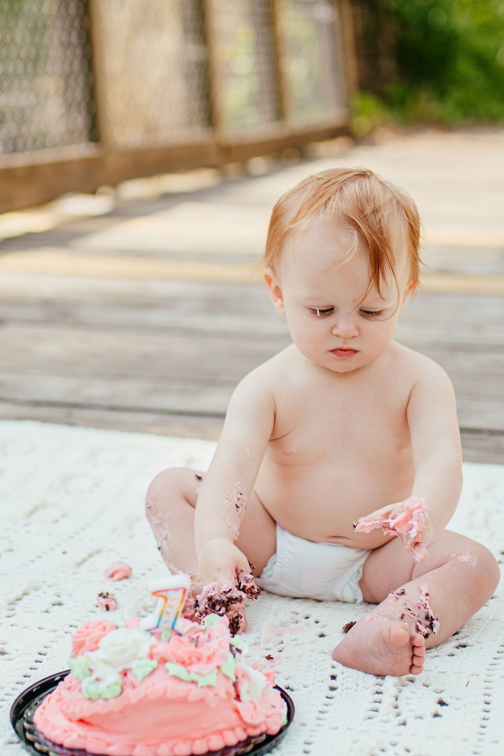 Toddler sitting on a blanket, looking at a pink frosted cake with frosting on hands and feet.