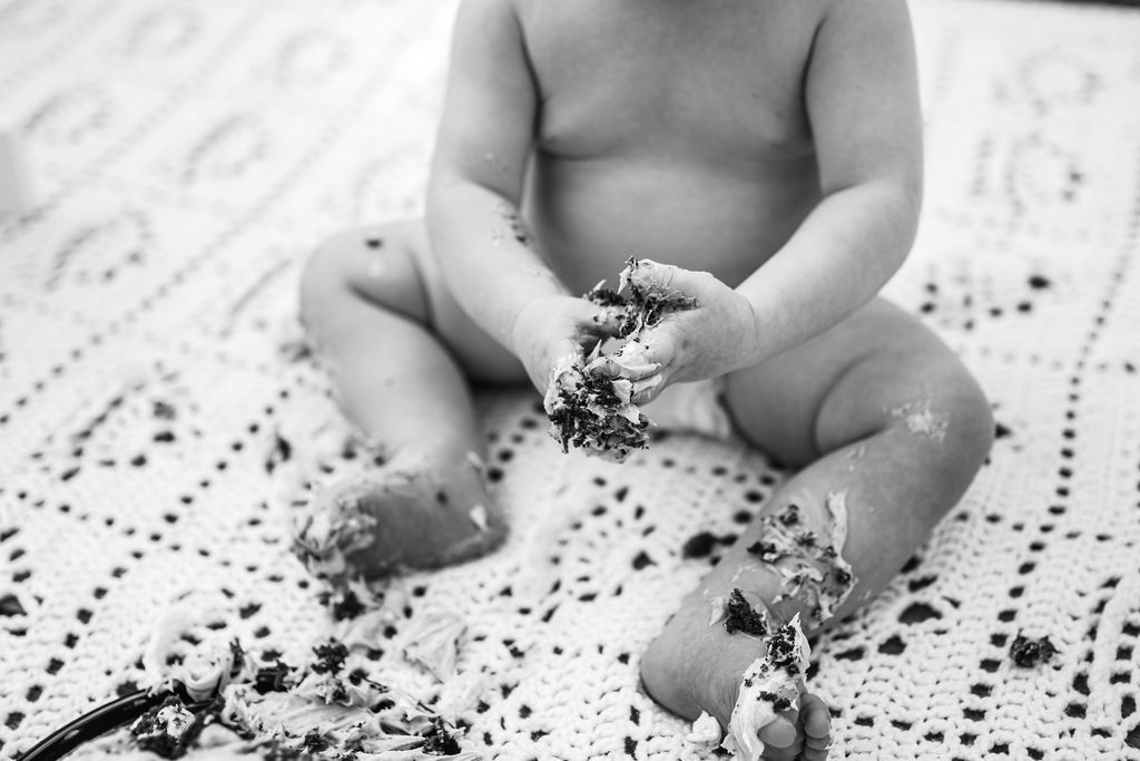 Baby sitting on a patterned blanket, playing with scattered grains or crumbs.