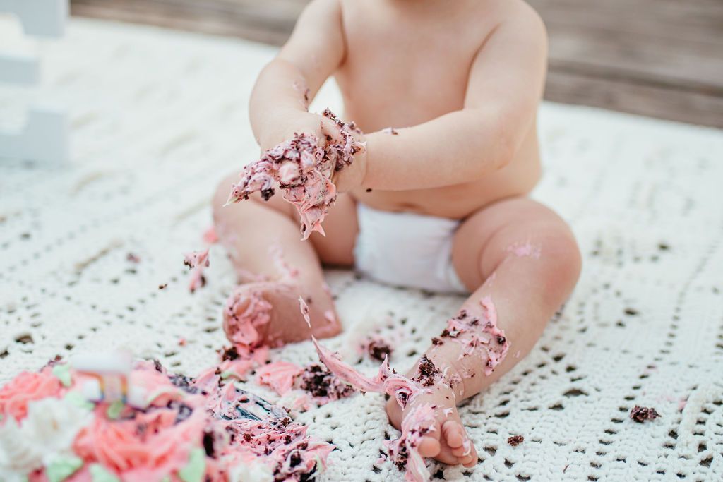 Baby sitting on a white blanket, covered in pink flower petals and looking at them with hands and feet.