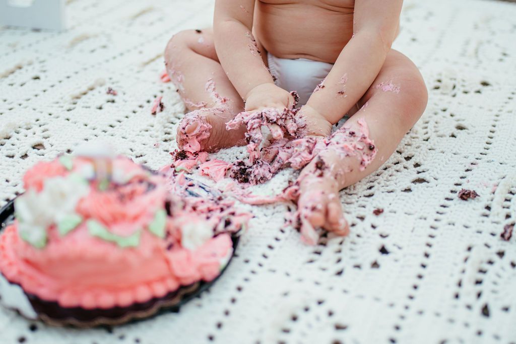 Toddler hands covered in pink frosting, next to a pink frosted cupcake on a lace tablecloth