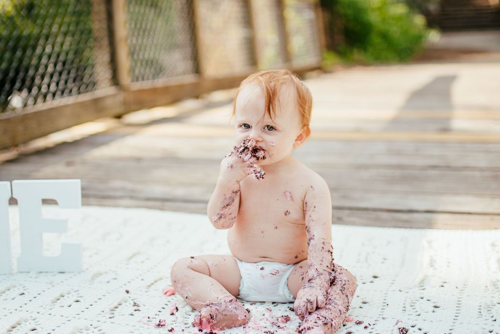 Toddler sitting on a white mat, playing with mud or dirt in an outdoor path area with a fence and greenery.