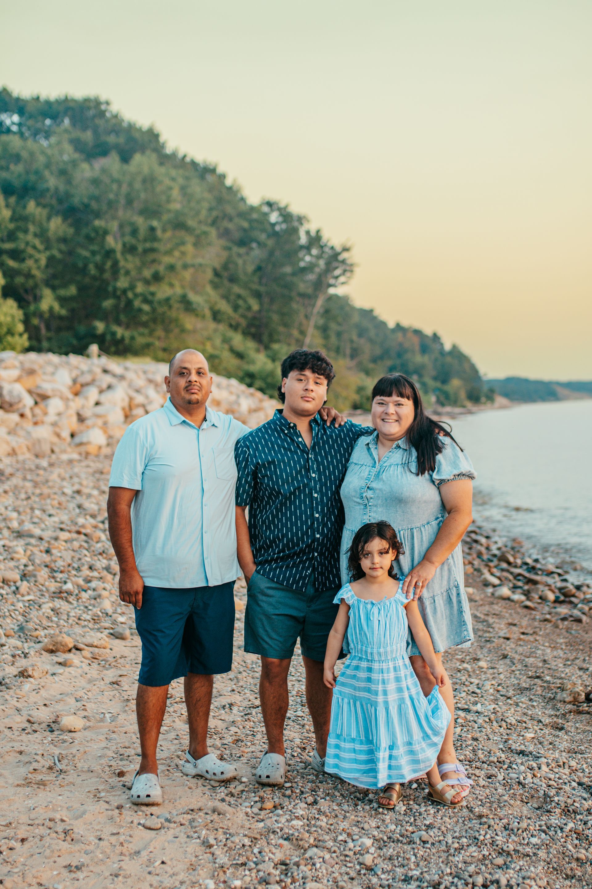 Family of four standing on a rocky beach at sunset, with trees along the shore and calm water nearby