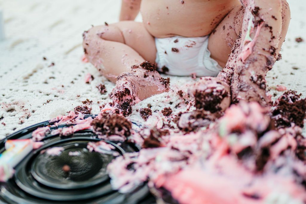 Baby covered in cake frosting, with a spilled cake on a table.