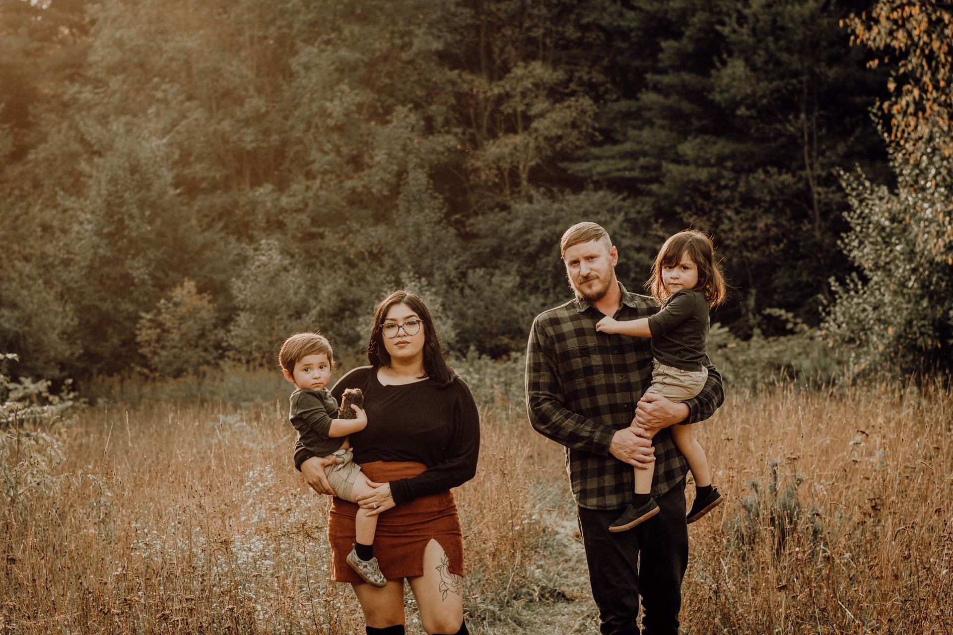 Family of four standing in a grassy field, holding children, with a forest background.