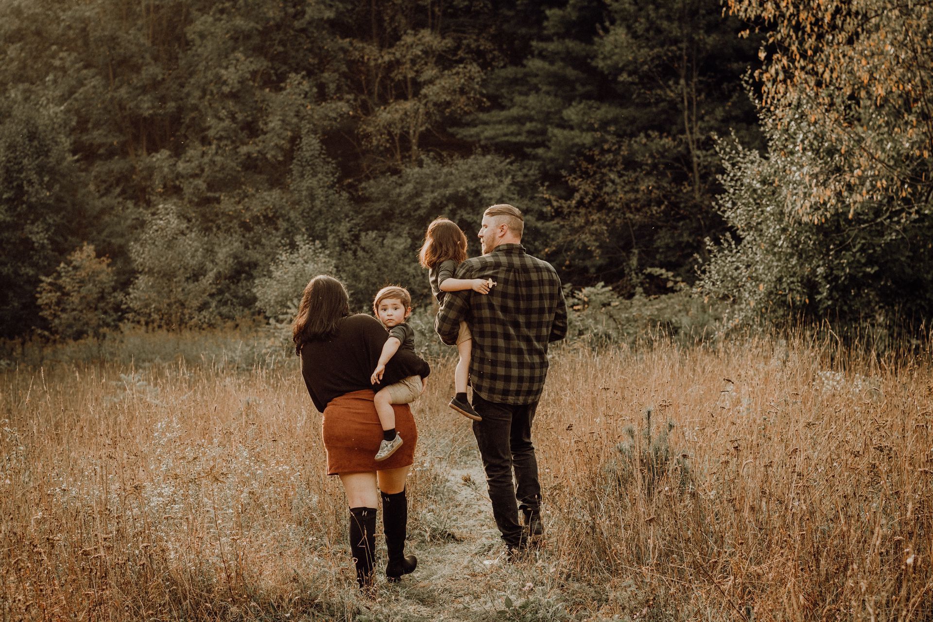 Family walking through a grassy field toward a wooded area, with one child being carried.