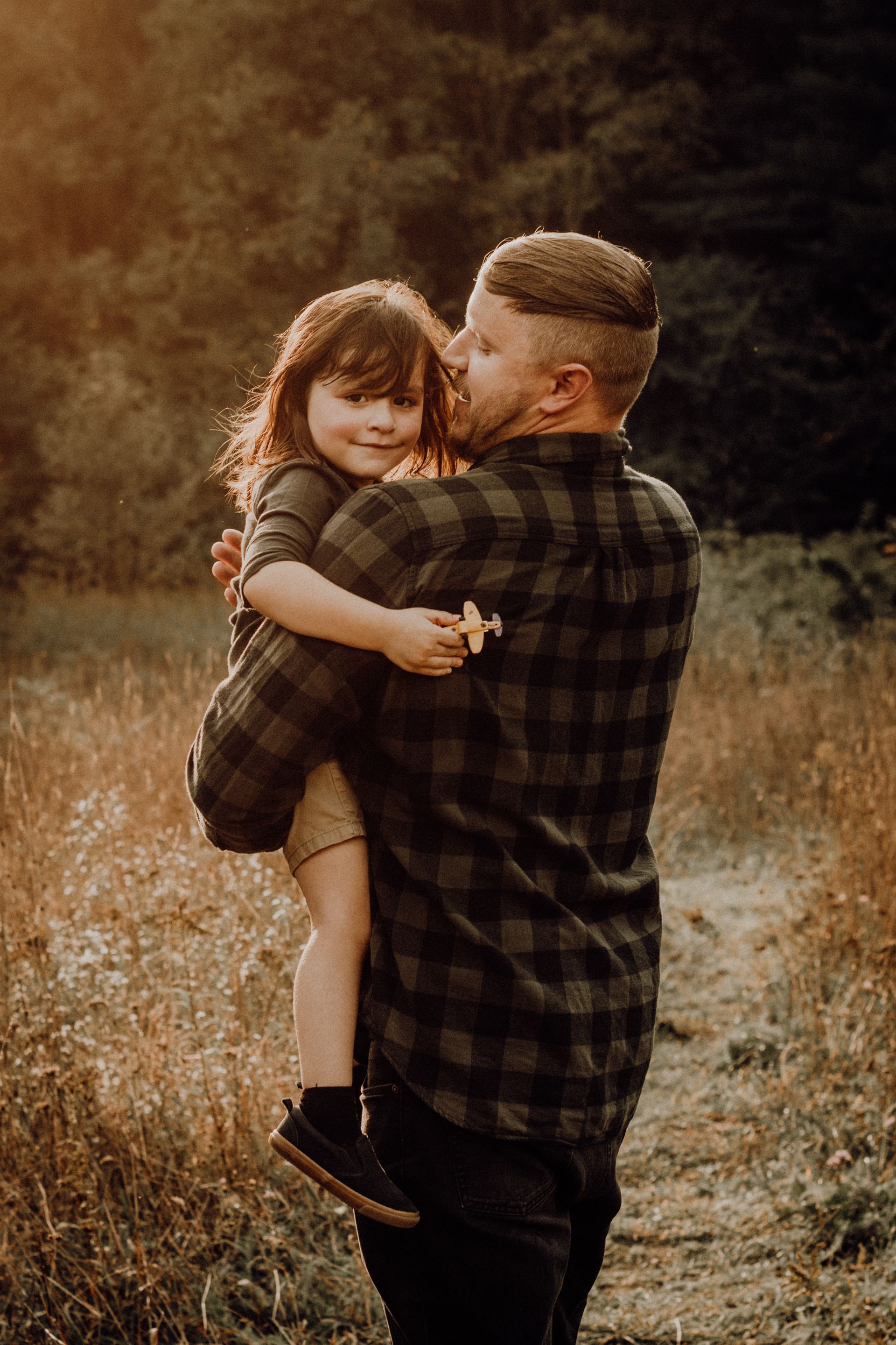Adult holding a smiling child in a golden field, both wearing dark plaid shirts.