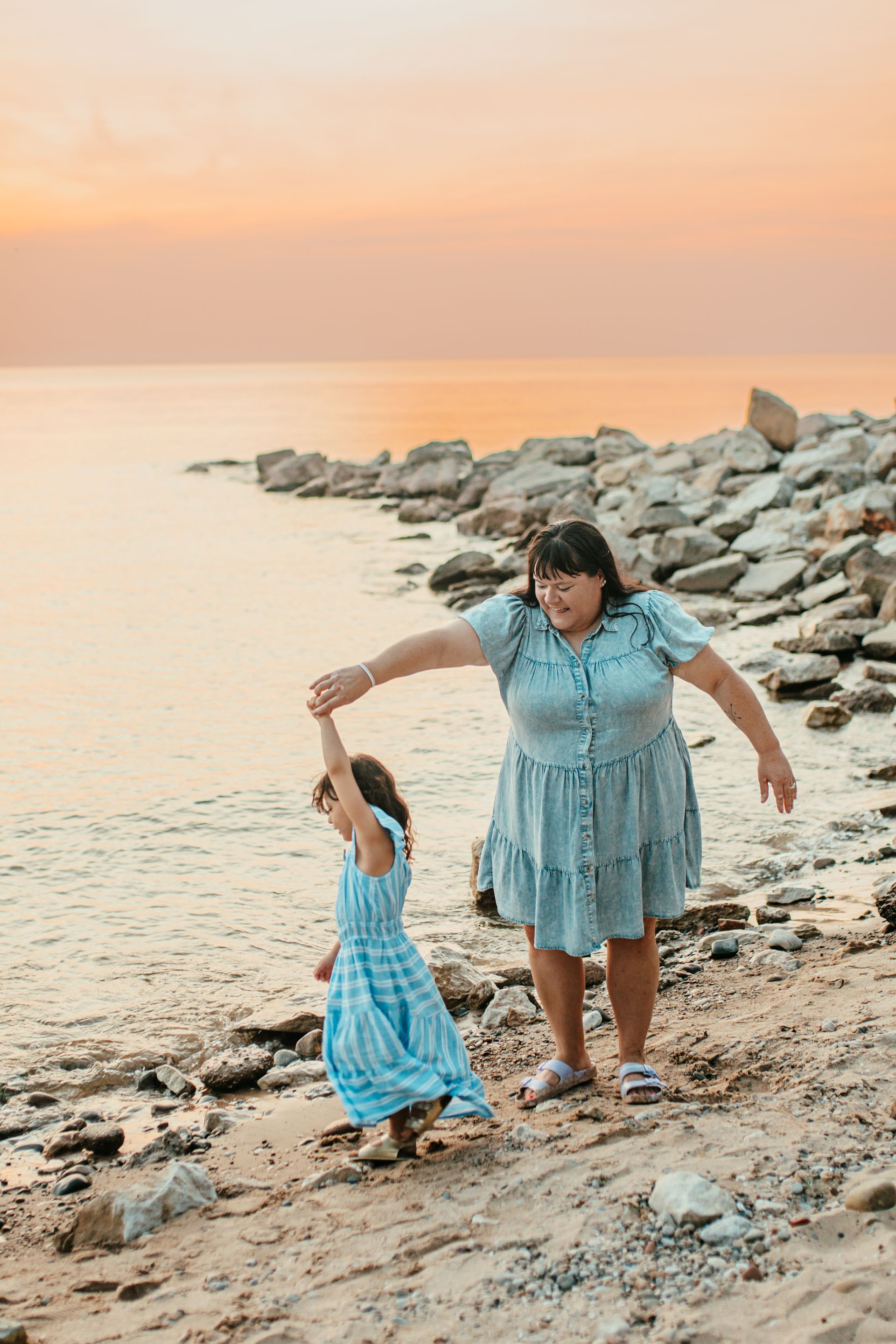 Adult and child holding hands on a rocky beach at sunset, wearing matching blue dresses