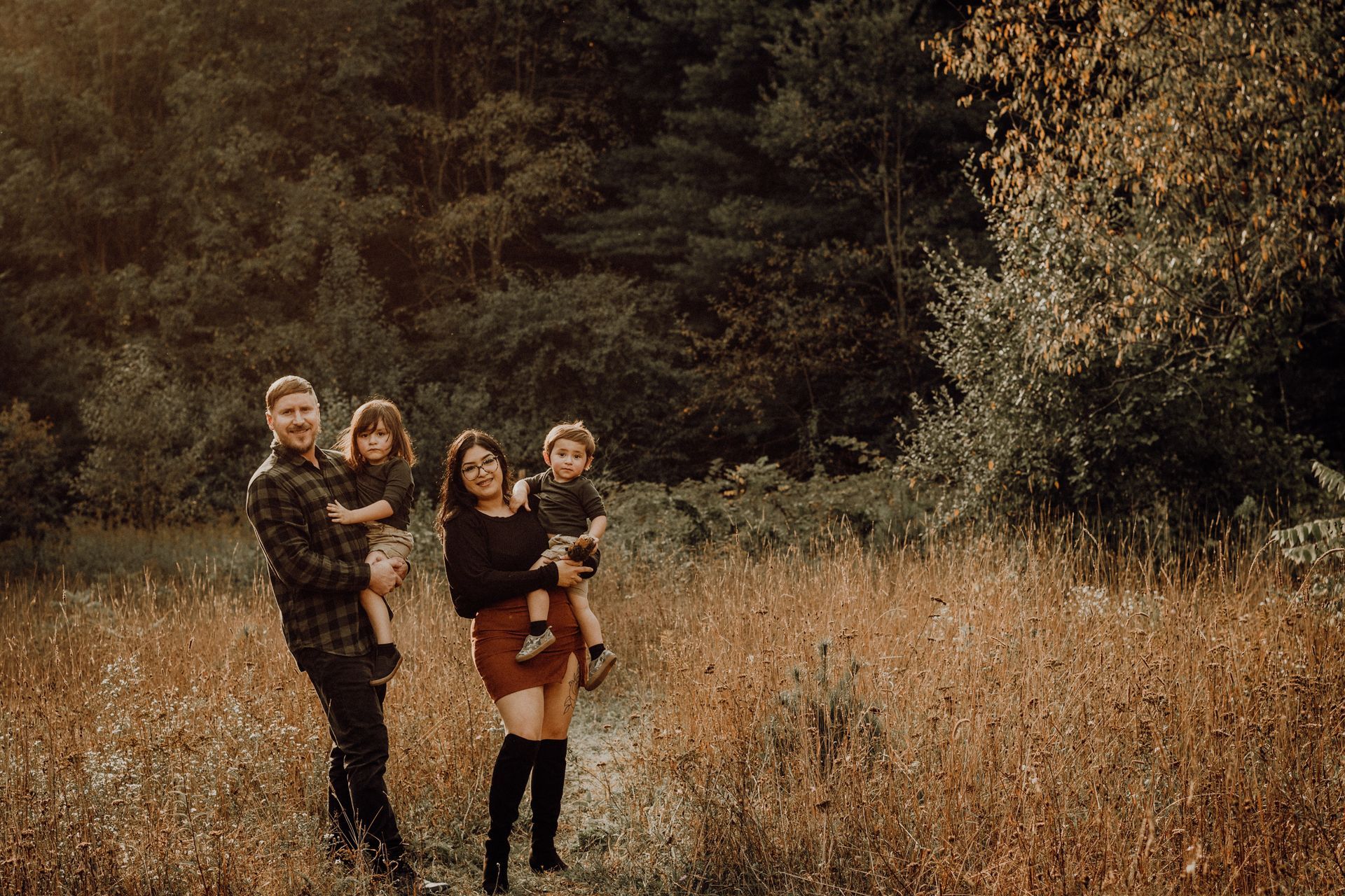 Four people standing in a grassy field with a dark forest backdrop, two holding children.