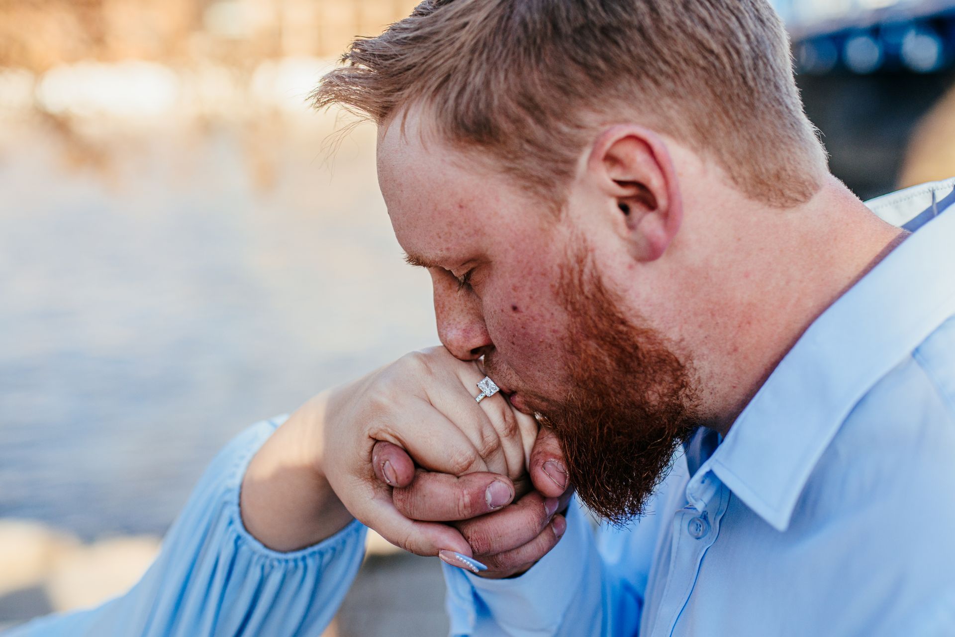 Man kissing another person’s hand outdoors near water, with a soft blurred background