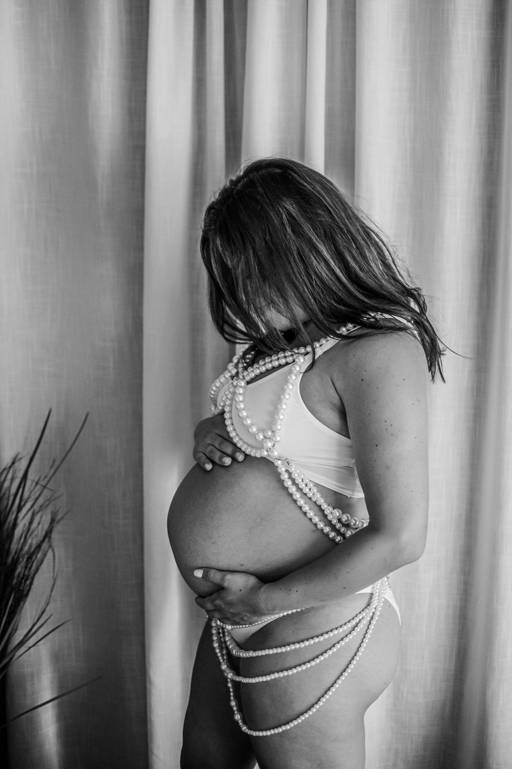 Pregnant woman in lingerie poses in profile against a curtain backdrop.