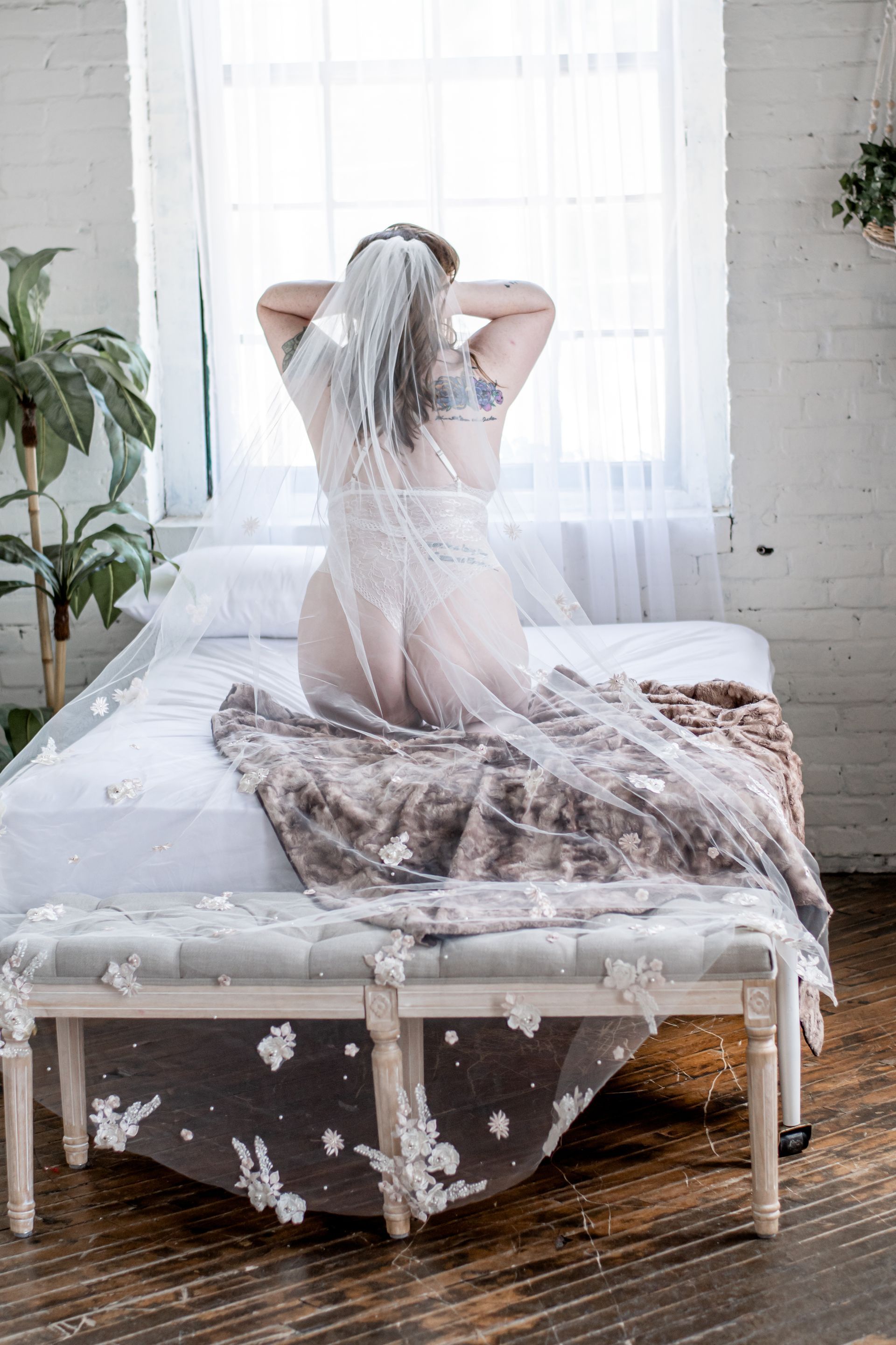 Woman sitting on a bed near a bright window, adjusting a sheer white veil in a softly lit room
