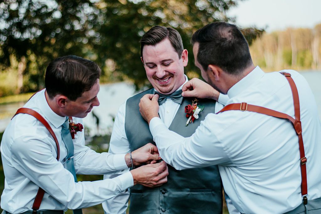 Three men in dress shirts and suspenders adjusting a groom’s vest outdoors by water